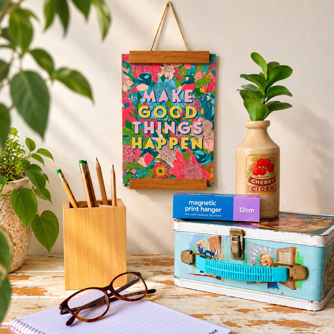A colorful wall-mounted print hanger with the phrase “MAKE GOOD THINGS HAPPEN” in bold multicolor lettering, styled on a rustic wooden table with a notebook, glasses, pencils, a vintage suitcase, and plants.