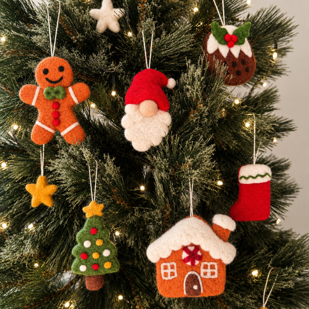 A collection of handmade felt Christmas ornaments hanging on a decorated green Christmas tree with warm white lights. The ornaments include a gingerbread man, Santa's beard, a pudding, a stocking, a Christmas tree, a gingerbread house, and a star.