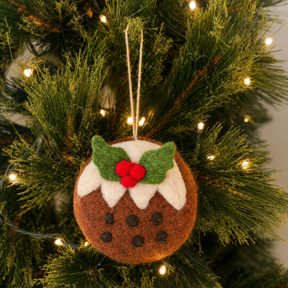 A brown felt Christmas pudding ornament with green leaves, white frosting, and red berries hanging on a decorated Christmas tree with warm string lights.