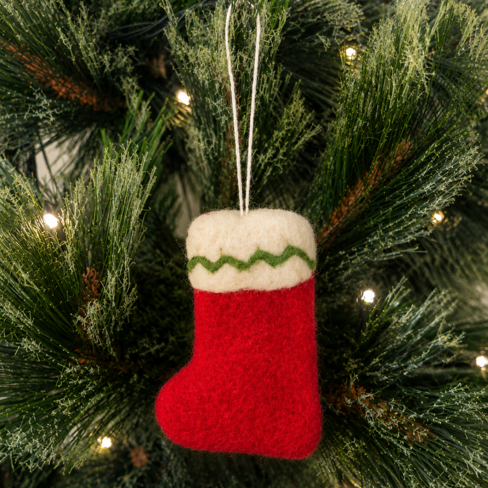 A red felt Christmas stocking ornament with a white cuff and green zigzag trim, hanging on a green Christmas tree with warm string lights.