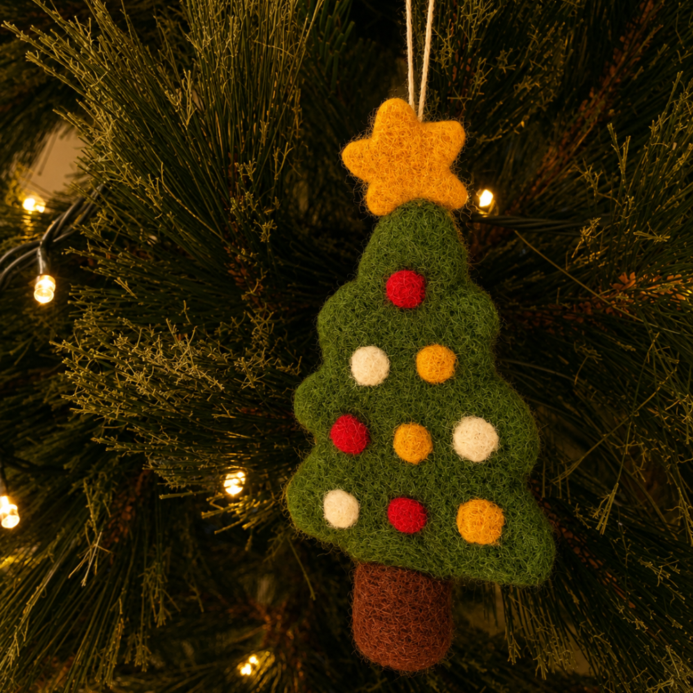 A felt Christmas tree ornament hanging on a pine tree branch with warm string lights. The ornament is dark green with a yellow star on top, decorated with red, yellow, and white felt pom-poms, and has a brown felt trunk.