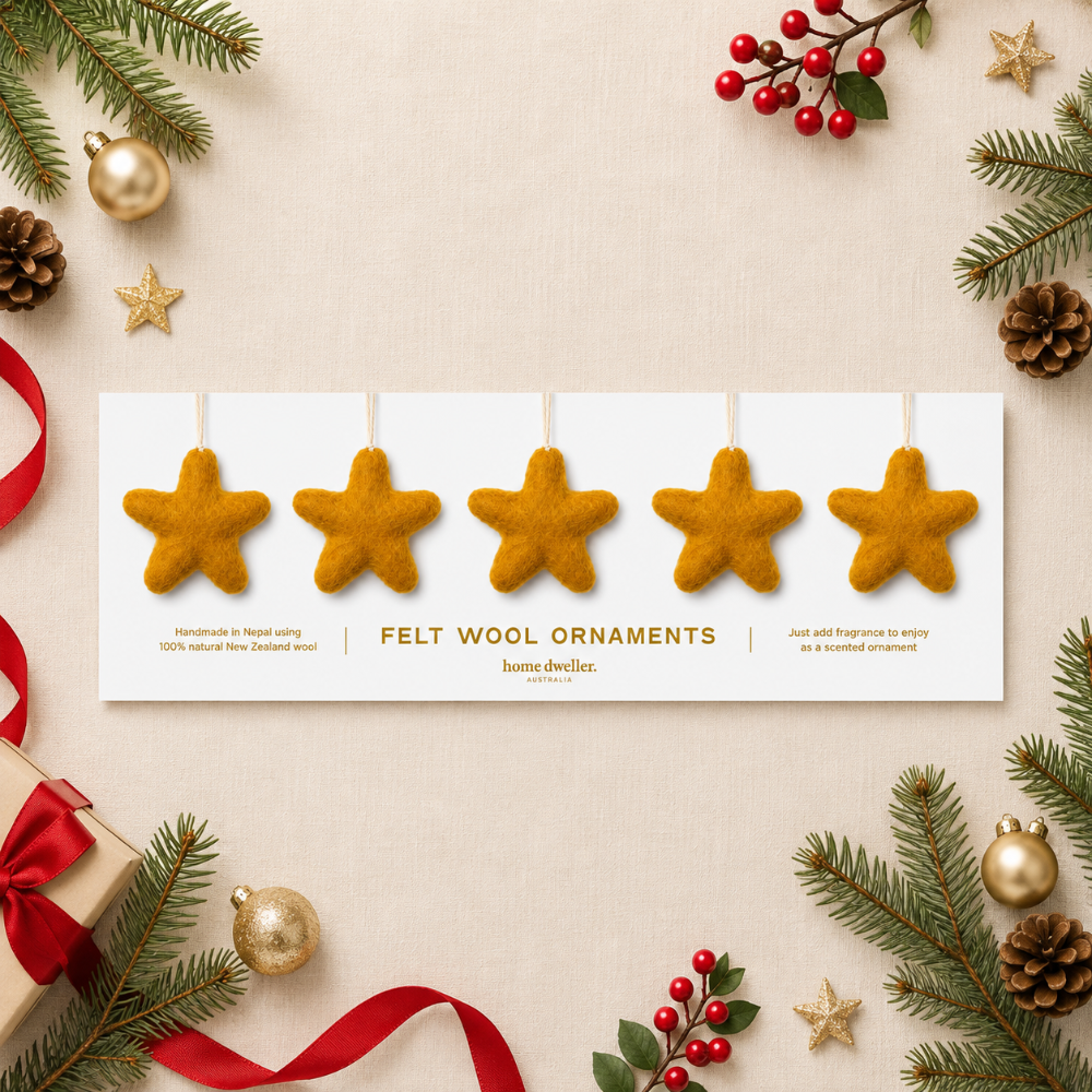 A flat-lay product presentation of five felt wool star ornaments on a white card, surrounded by festive Christmas decorations including pine branches, gold ornaments, red berries, and a wrapped gift on a beige linen background.