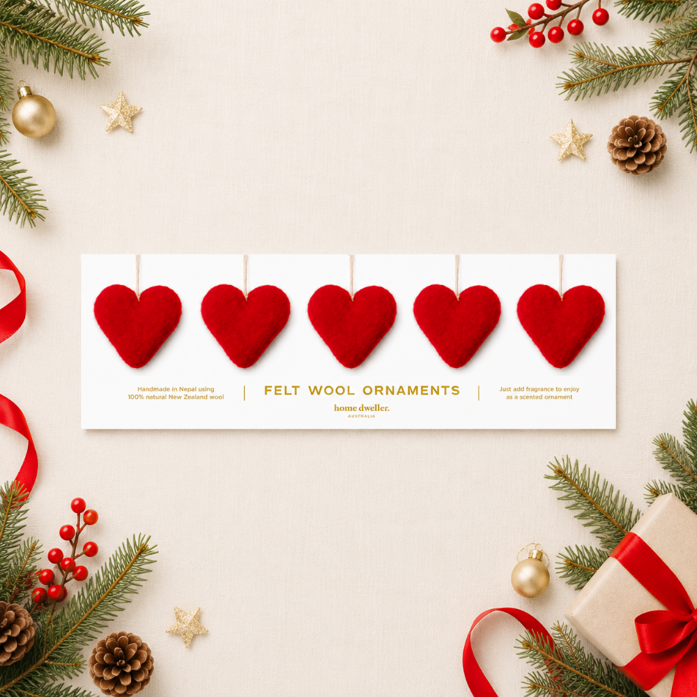 A flat-lay product shot of a white card displaying four red felt wool ornaments, surrounded by festive Christmas decorations including pine branches, red berries, gold stars, pinecones, and a wrapped gift box on a light beige background.