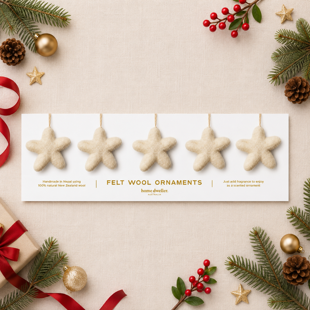 Five beige felt wool star ornaments hang from a white display card on a light beige fabric background, surrounded by holiday decorations including pine branches, red berries, gold ornaments, and a wrapped gift.