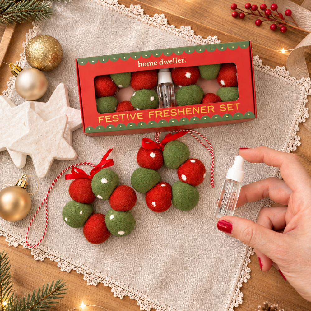 A festive home fragrance gift set displayed on a lace-trimmed beige cloth over a wooden surface. The set includes a red box labeled 'home dweller' and 'FESTIVE FRESHENER SET' with small bottles and red-and-green felt holiday wreath ornaments.