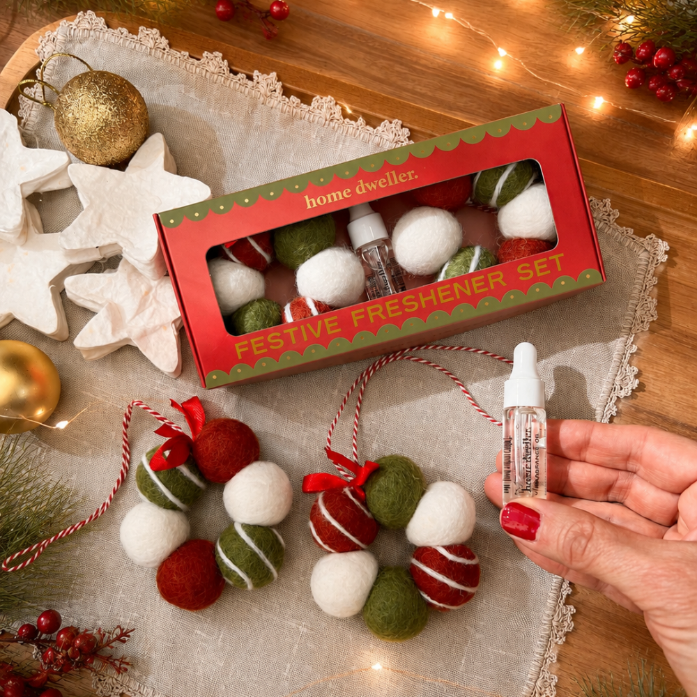 A hand-dyed Christmas-themed home fragrance set displayed on a rustic wooden table with holiday decorations. The set includes a red and green box of 'home dweller' FesToe Freshener Set, two felt wool ornaments in red, white, and green, and a small hand-painted glass dropper bottle being held by a hand with red nail polish.