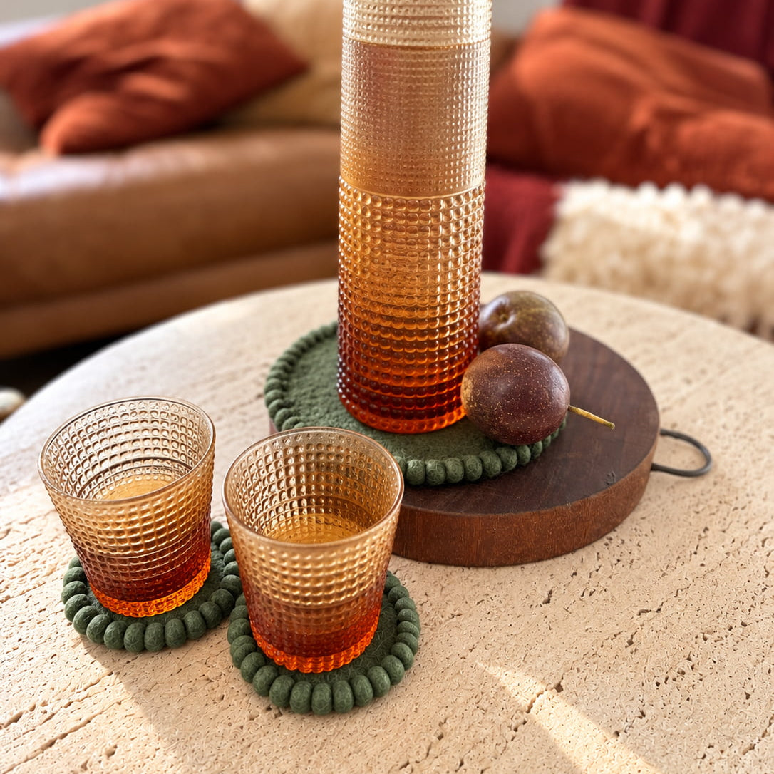 A set of amber-colored textured glassware, including a tall pitcher and two tumblers, arranged on a round table in a cozy living room setting.