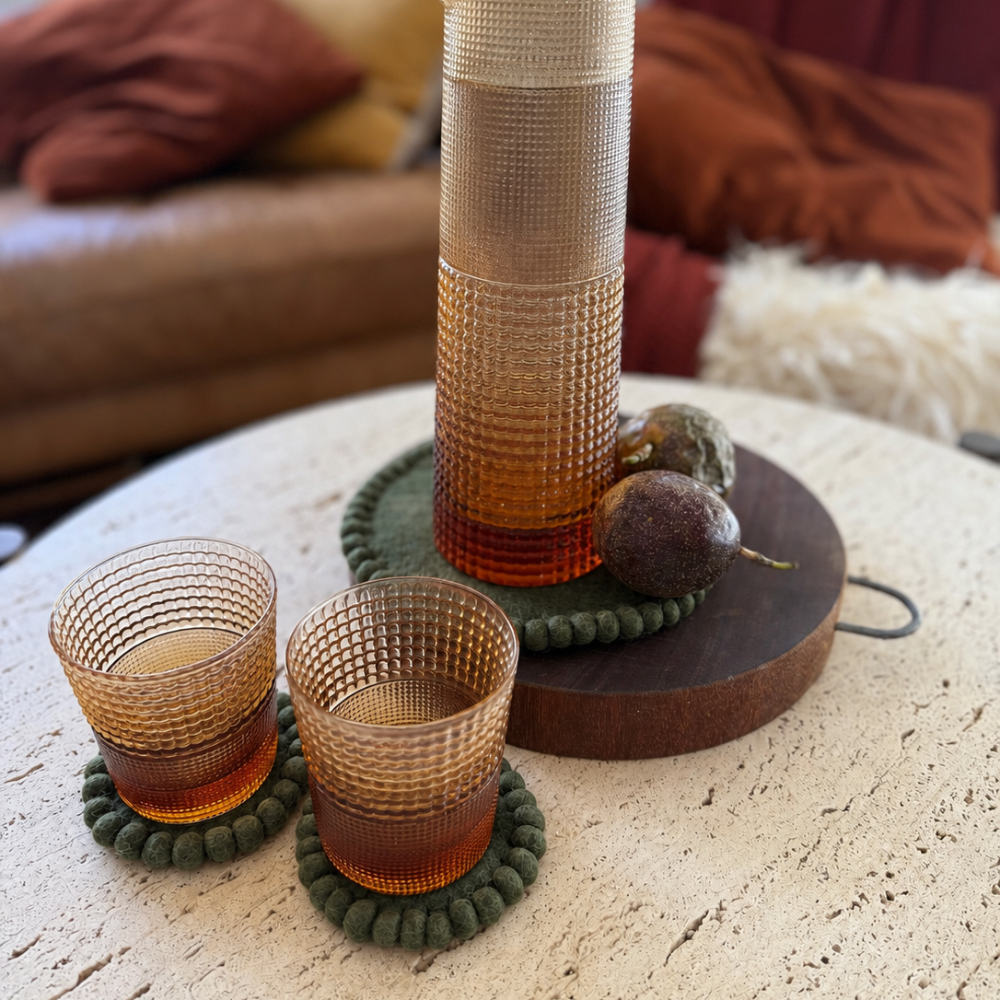 A set of amber glass tableware including a tall carafe and two matching textured tumblers with dark green beaded coasters, arranged on a round table in a cozy living room setting.