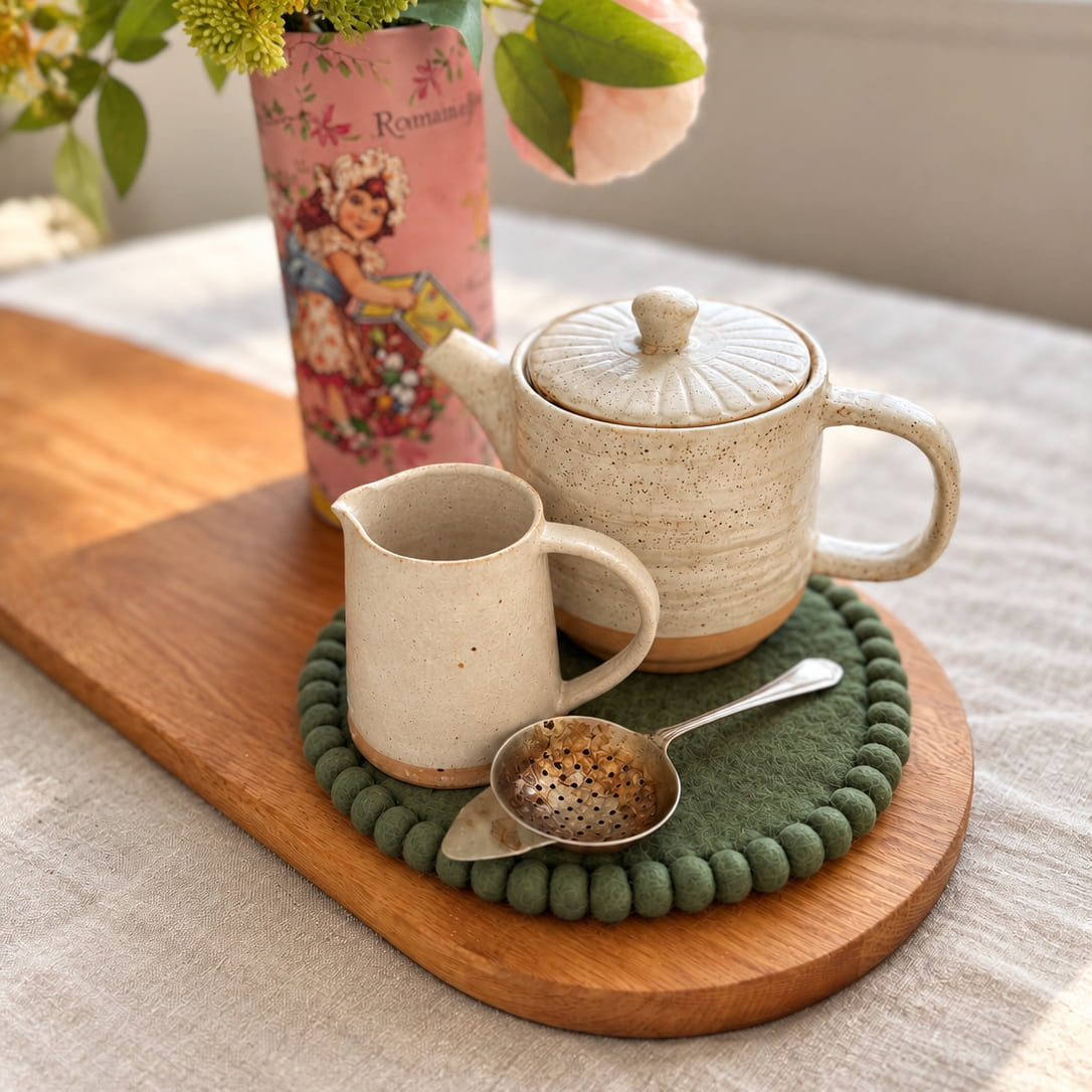 A ceramic tea set arranged on a wooden tray with a green pom-pom trivet, including a speckled teapot, cream pitcher, tea infuser, and matching coaster. A pink floral vase with flowers sits behind on a light tablecloth near a window.