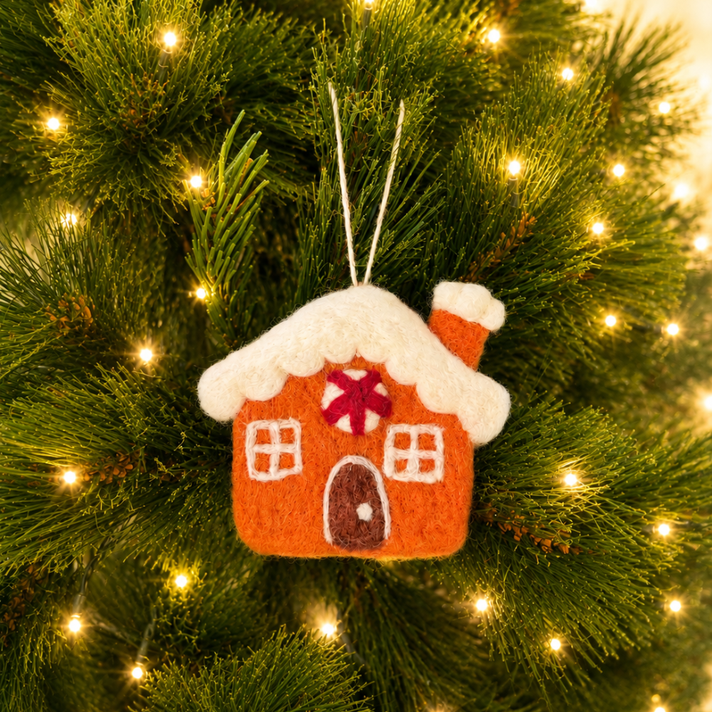 A small felt Christmas house ornament hanging on a green Christmas tree with warm white string lights.