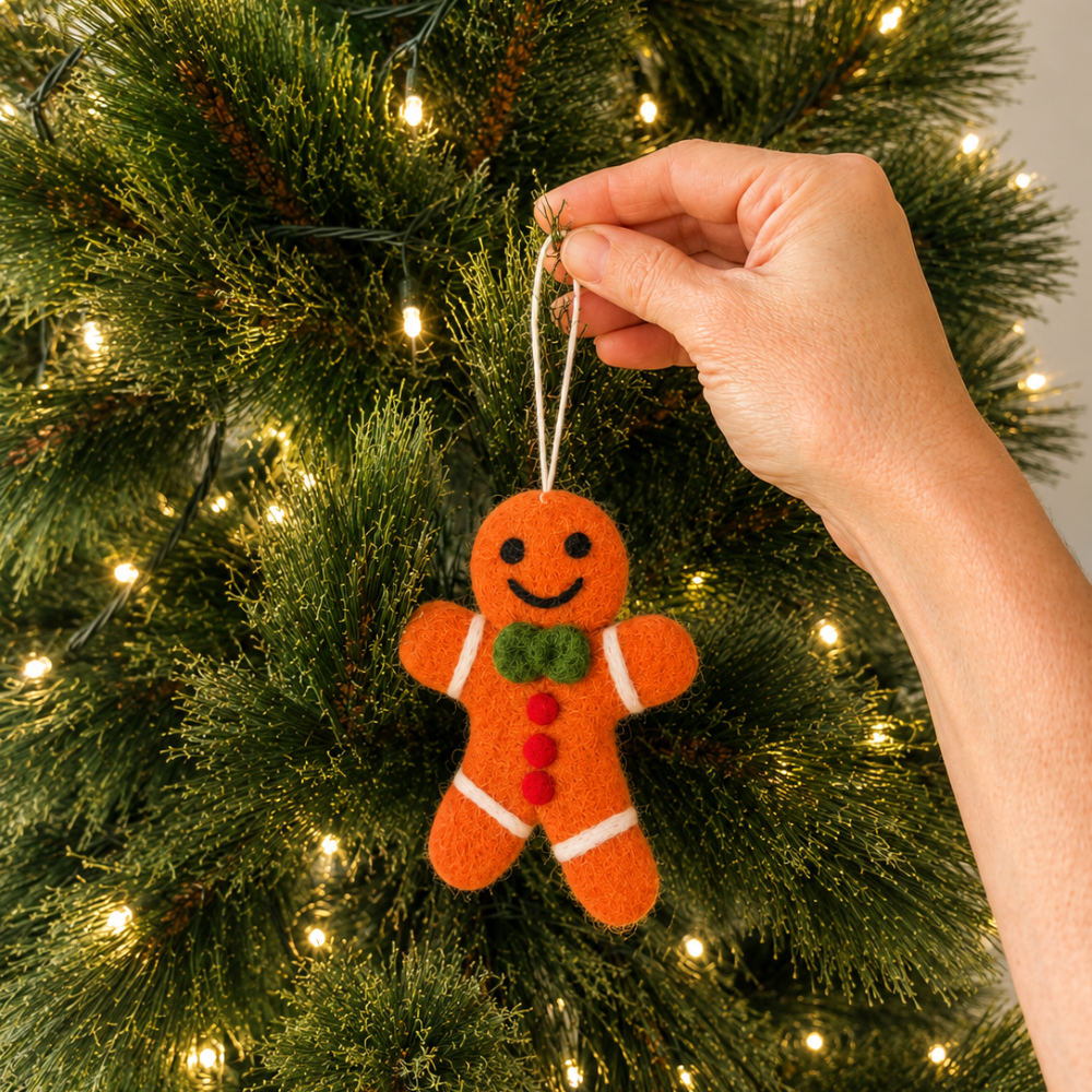 A hand hanging an orange felt gingerbread man Christmas ornament with a green bow tie and red buttons from a lit green Christmas tree.