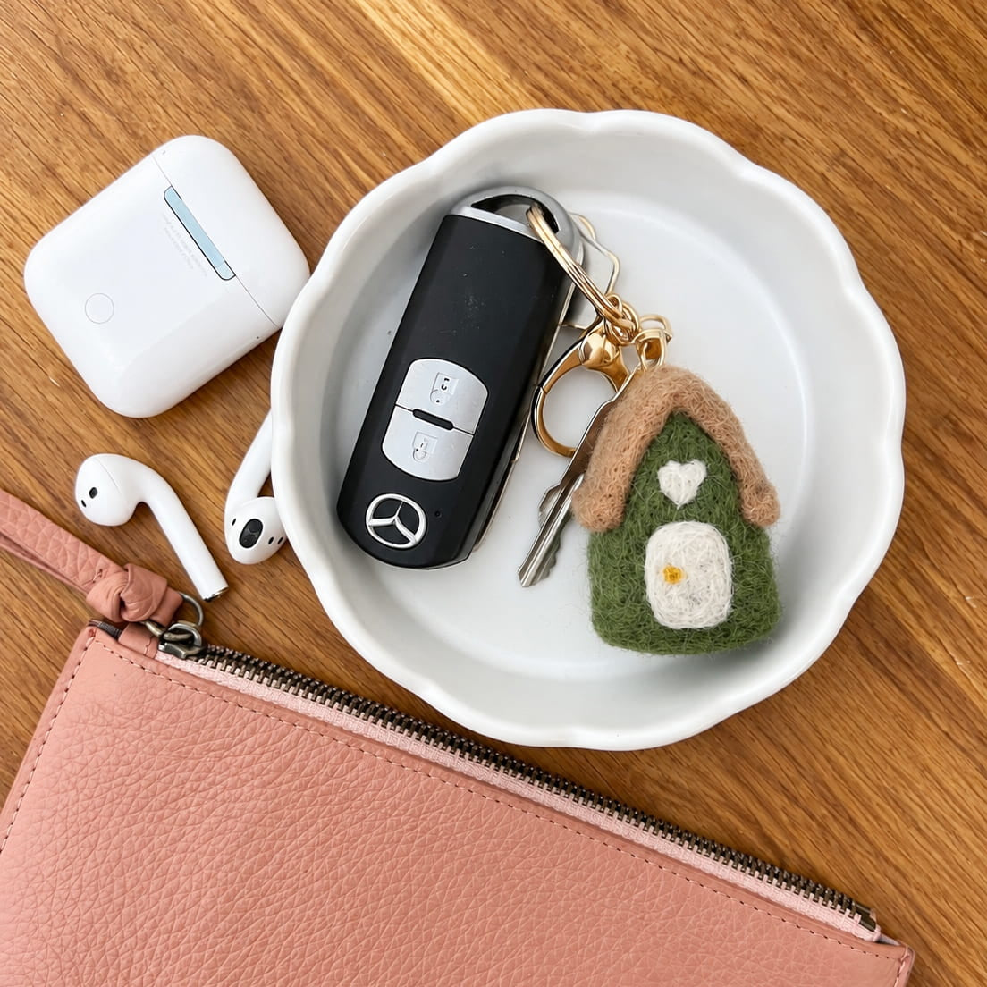 A styled flat-lay on a wooden table featuring a pink pebbled leather zip pouch, a white ceramic dish holding a Mercedes key fob and a green felt house keychain, alongside white wireless earbuds in their charging case.