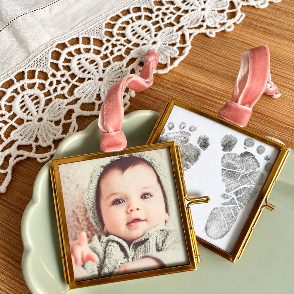 Two small gold-framed keepsake photo frames with pink ribbon loops, displayed on a pale green dish over a wooden surface with a white lace cloth. One frame shows a baby portrait, and the other shows a baby footprint print on white paper.