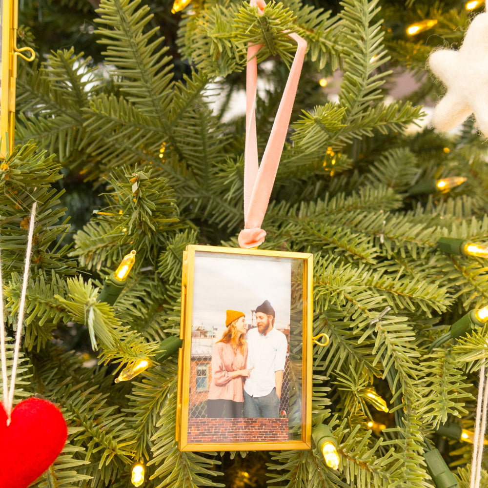 A gold-framed photo ornament hanging on a green Christmas tree with warm string lights. The ornament contains a printed photo of a couple and hangs from a pink ribbon.