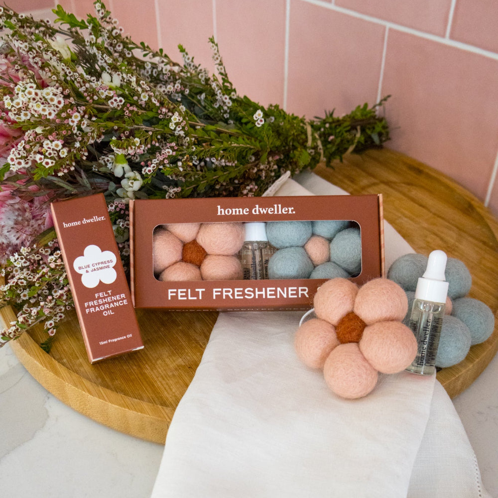 A home fragrance gift set from home dweller, featuring a box of felt freshener balls, a bottle of essential oil, and a flower-shaped felt diffuser, styled on a wooden board with a white cloth against a pink tiled background.