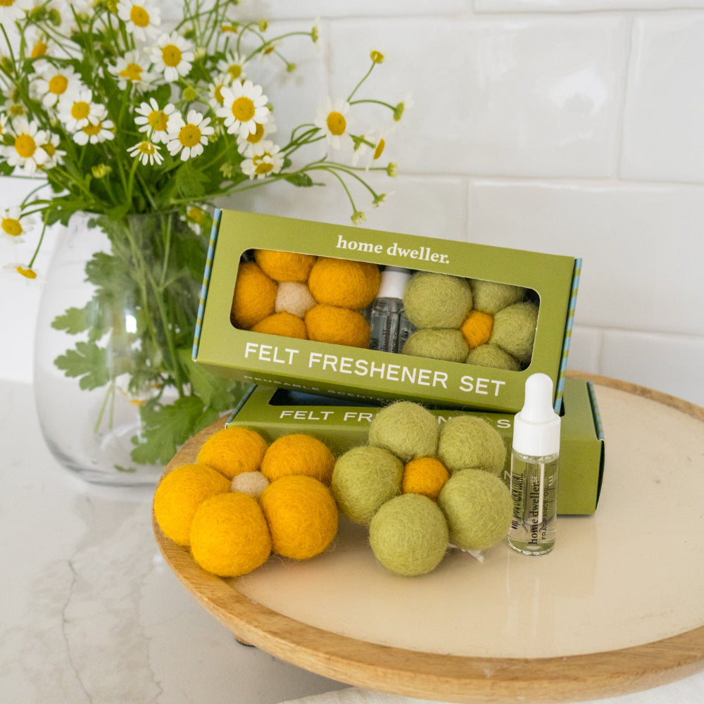 A felt freshener set by home dweller, featuring green and orange felt balls in a matching olive green box, displayed on a wooden tray next to a small dropper bottle. A glass vase of daisies sits in the background against a white subway tile wall.