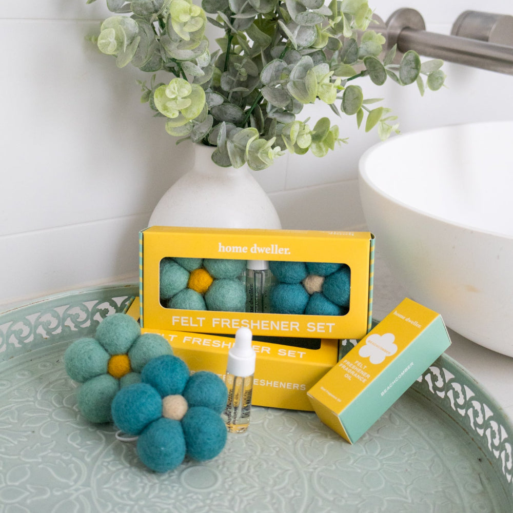 A home fragrance set displayed on a decorative tray, featuring a yellow box labeled 'home duffel' containing felt freshener beads, a small bottle, and a mint green box, styled with a white vase of eucalyptus and a white sink in the background.