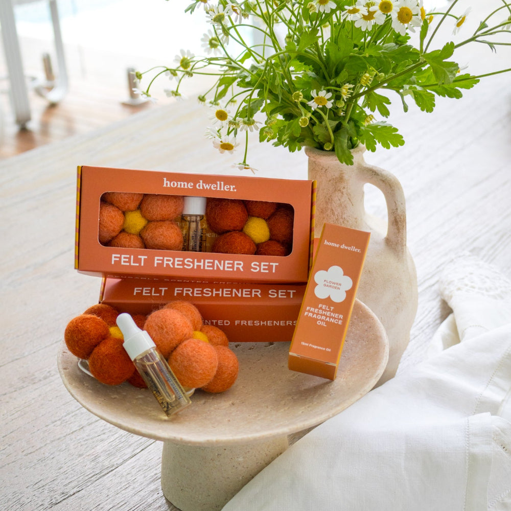 A home fragrance product set displayed on a ceramic pedestal tray on a light wood floor, featuring orange felt diffusers, a small amber bottle, and an orange box labeled 'home dvelices Felt Freshener Set,' with a vase of daisy flowers and a white cloth nearby.
