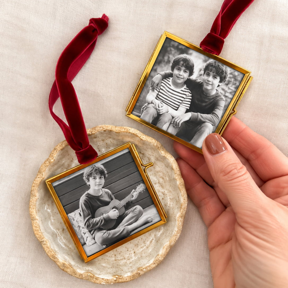 Two square gold-framed photo ornaments with red velvet hanging ribbons are displayed on a light fabric surface. One ornament rests on a round textured dish, and the other is held by a hand.