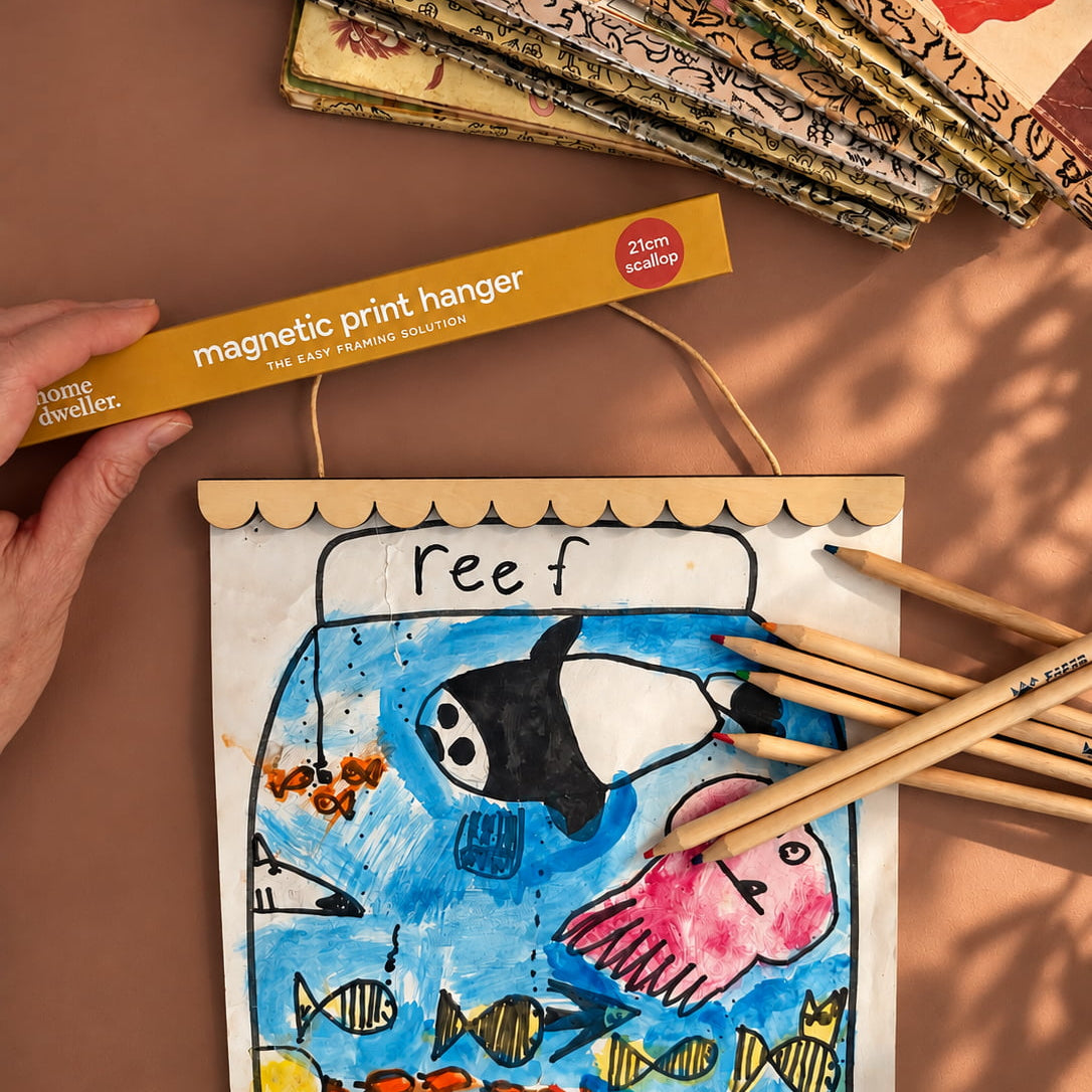 A magnetic print hanger holding a colorful children's drawing of an underwater reef scene, shown with wooden pencils and a stack of printed samples on a warm brown surface. A hand holds the hanger packaging, and the scene includes visible shadows from outdoor light.