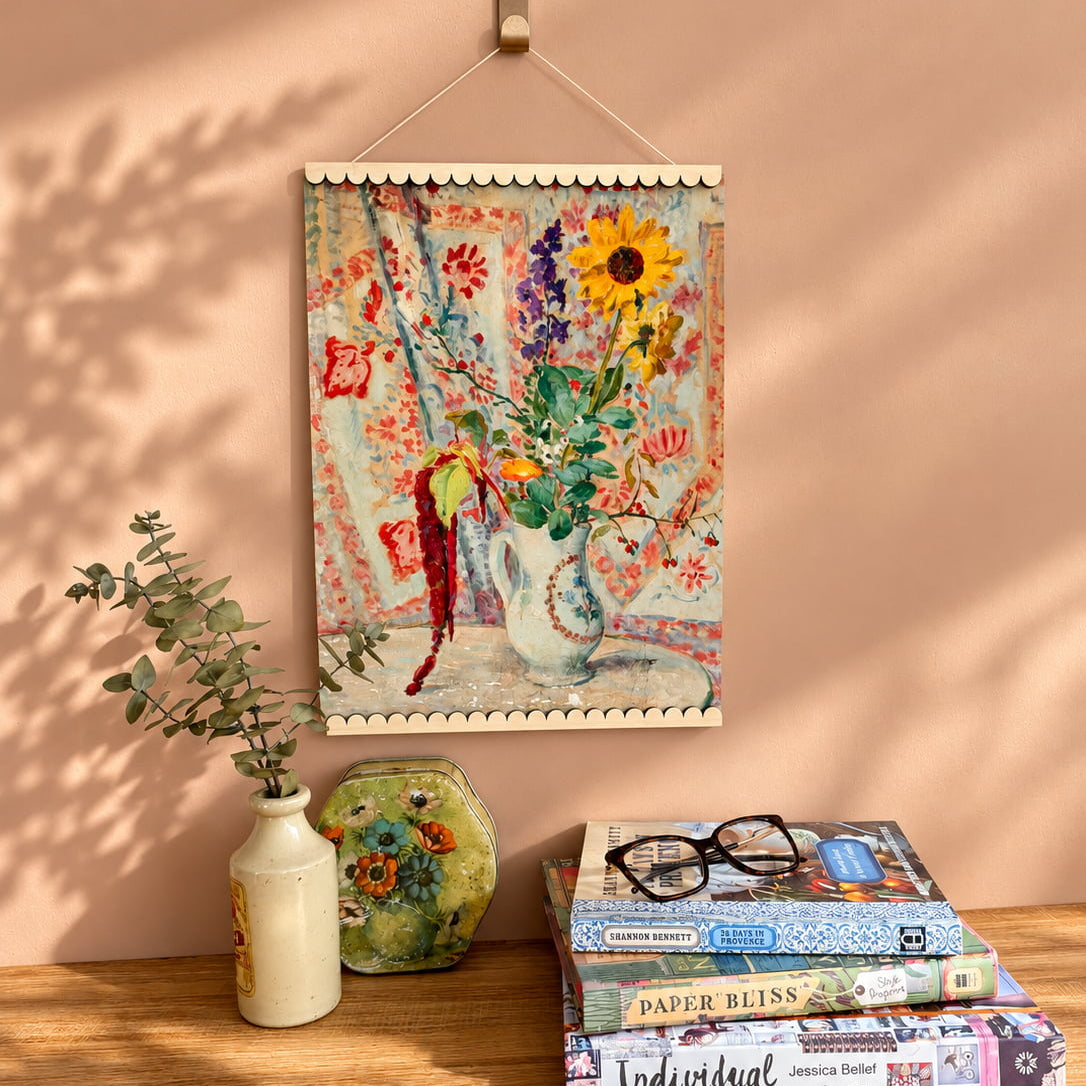 A colorful floral tapestry wall hanging with a sunflower design is displayed on a wooden shelf against a peach-colored wall with leaf shadows. The tapestry is styled with a small white vase, a green ceramic object, and a stack of books on a wooden table.