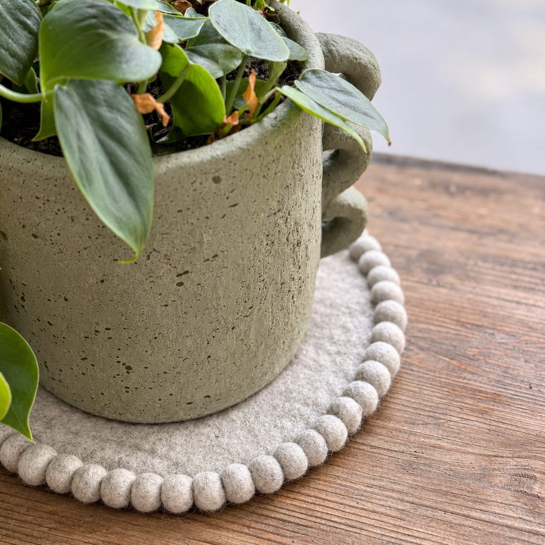 A green leafy plant in a speckled light gray concrete planter with sculptural side handles, placed on a round beige felt coaster with a beaded edge on a wooden table.