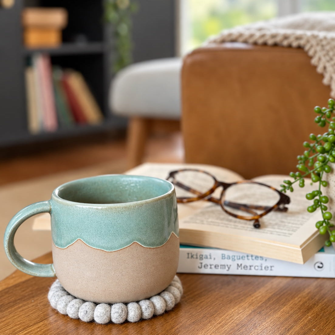 A ceramic mug with a teal and beige color scheme sits on a wooden table next to an open book and eyeglasses, with a cozy living room background.