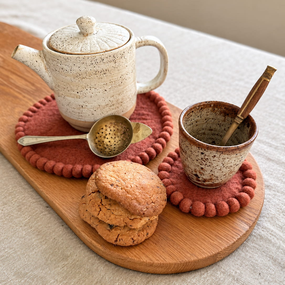 A ceramic tea set and matching cup arranged on a wooden serving board with cookies and a metal tea infuser, set on a light linen tablecloth.