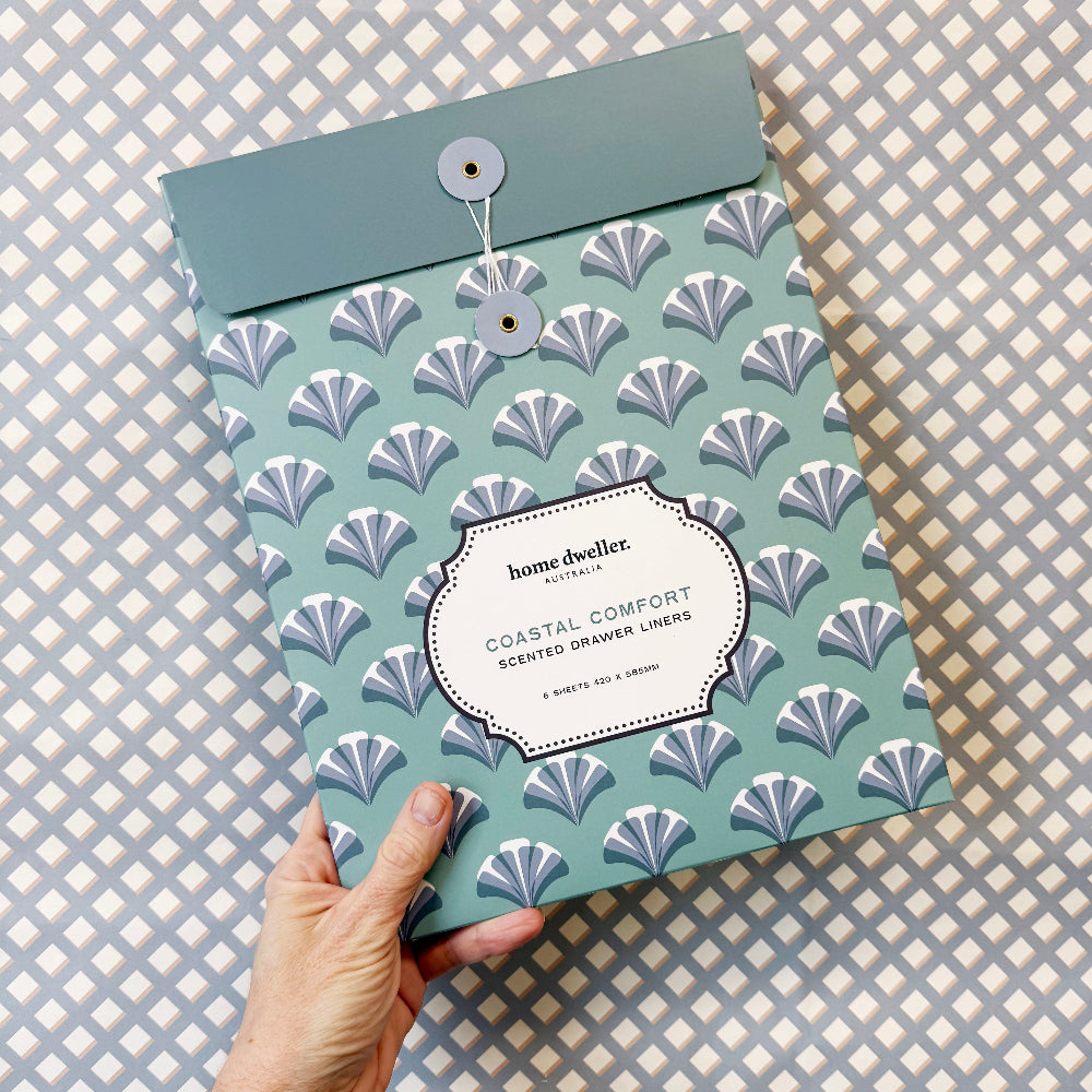 A hand holds a square envelope-style package for Home Dwellers Coastal Comfort Scented Flower Liners against a gray and white patterned background.